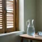 Les 6 meilleurs réflexes santé face à la 4e canicule 2026 Intérieur frais d’un appartement pendant une canicule, avec des volets fermés, une carafe d’eau et un thermomètre près d’une fenêtre.