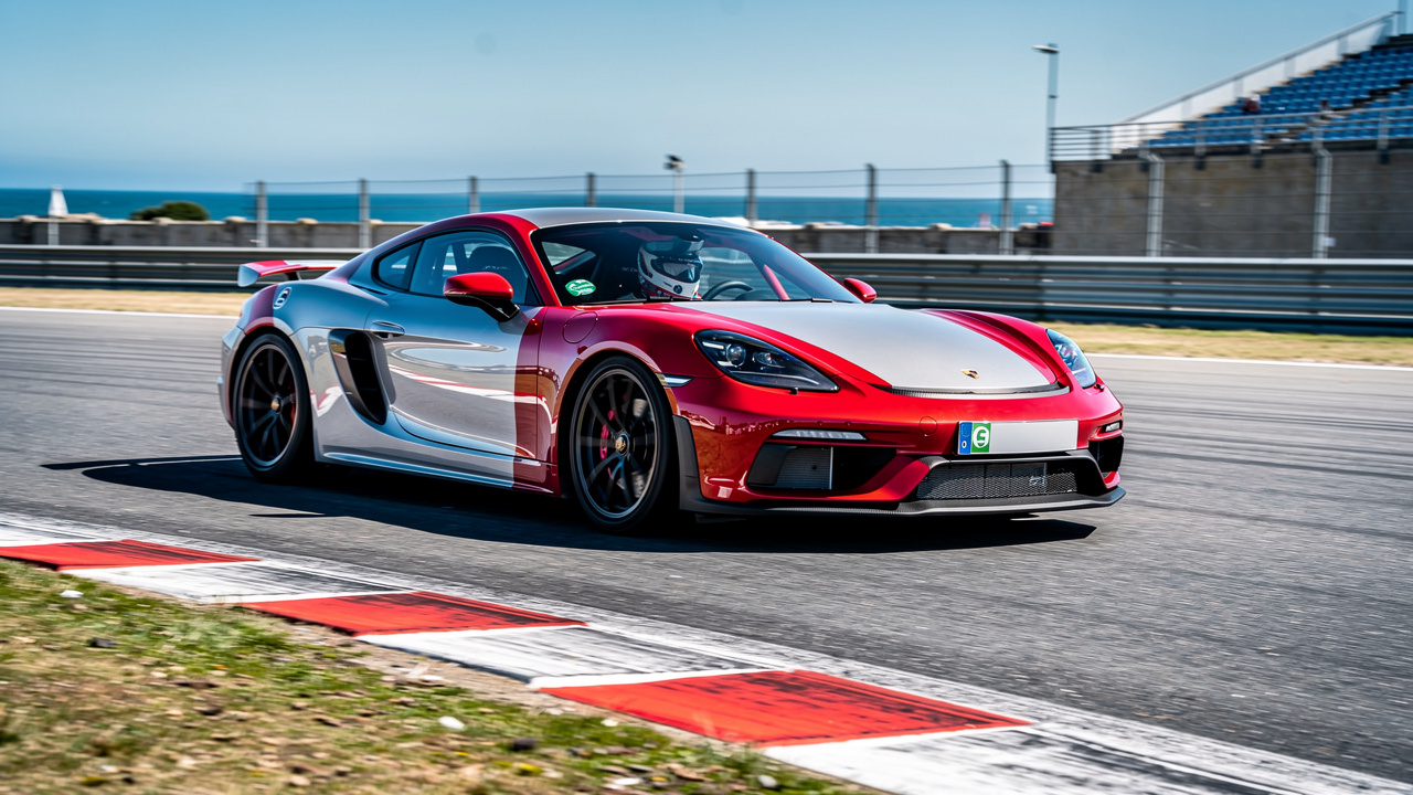 Une voiture de sport rouge et argentée conduite par un pilote casqué dans un virage d’un circuit breton, près de vibreurs rouges et blancs sous un ciel lumineux.