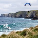 12 activités à sensations fortes dans le Finistère Panorama du Finistère montrant un surfeur dans les vagues, une parapentiste au-dessus de falaises sauvages et la côte bretonne baignée d’une lumière douce.